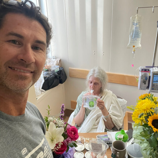 Marsha in the hospital during early recovery from a fractured vertebra, supported by her trainer and surrounded by flowers.